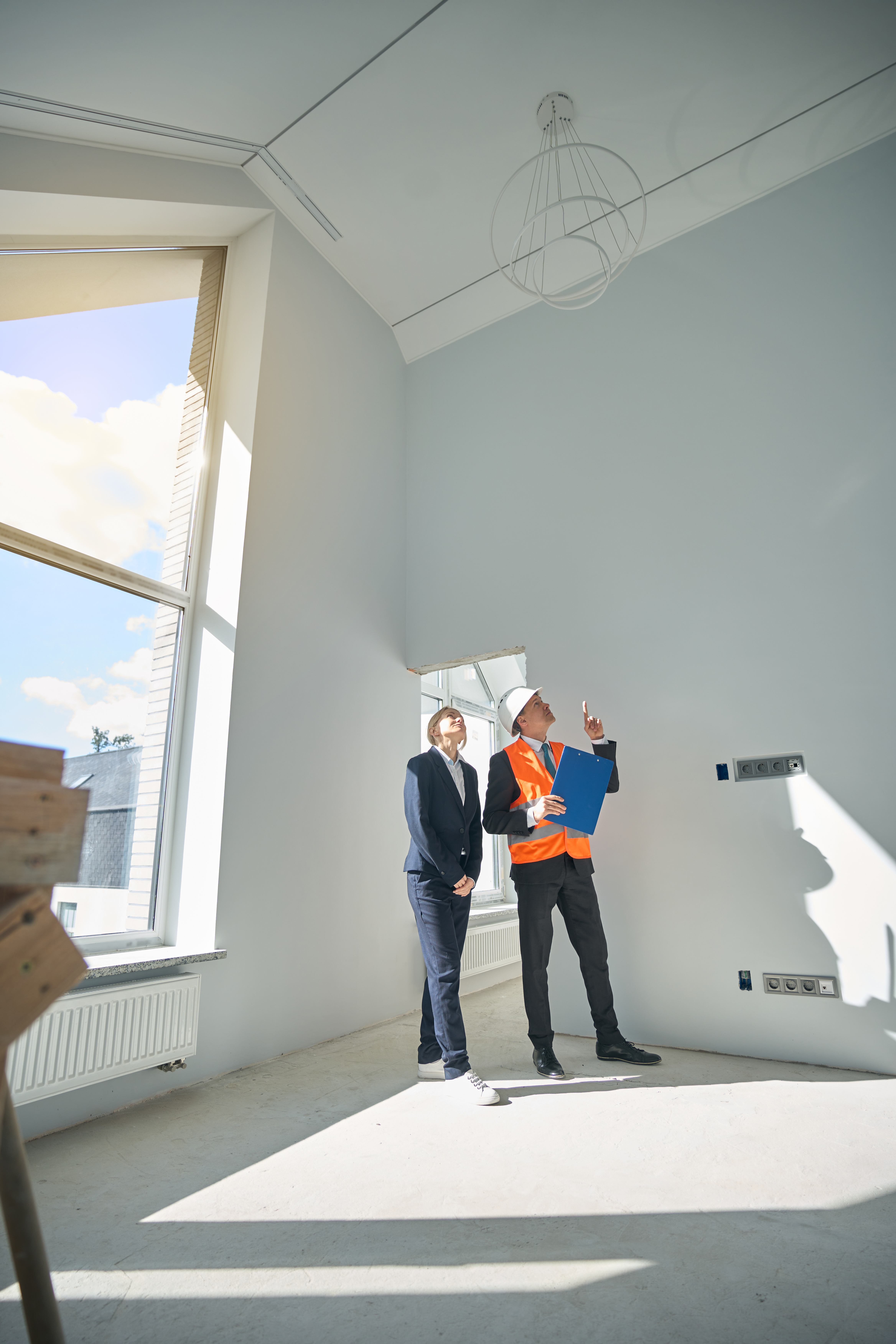 Male foreman pointing finger up while woman client looking at ceiling when both of them standing in empty room