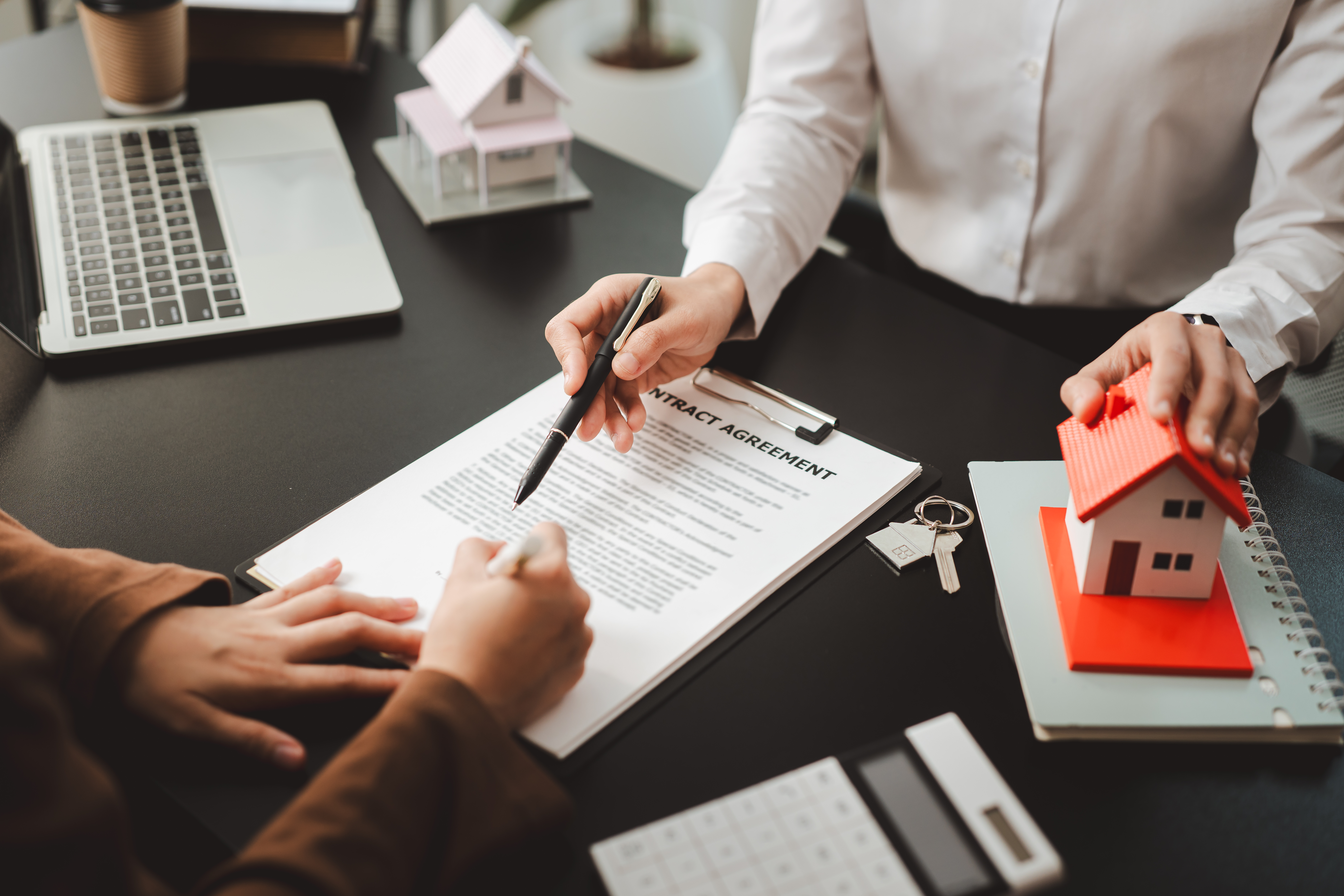 Two people closing a real estate deal at a desk, one signing a contract while the other holds a model house, highlighting homeownership, investment and financial planning